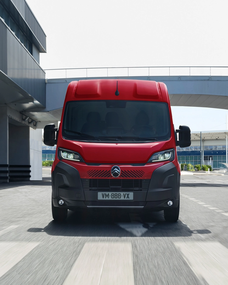 Front view of a red Citroën Relay van driving through a modern urban setting, highlighting robust design, practicality, and a strong road presence.