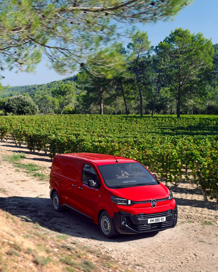 Three‑quarter front view of a red Citroën Dispatch driving on a rural road, showing robust design, driving comfort, versatility and confident presence.